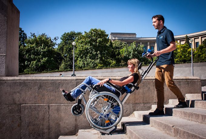 Jeune femme en fauteuil roulant descendant un escalier grâce à un monte-escaliers autonome manipulé par une tierce personne.