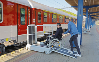 Un aidant fait monter une personne en fauteuil roulant sur la plateforme élévatrice mobile Pégase afin qu'il puisse accéder au train entré dans la gare
