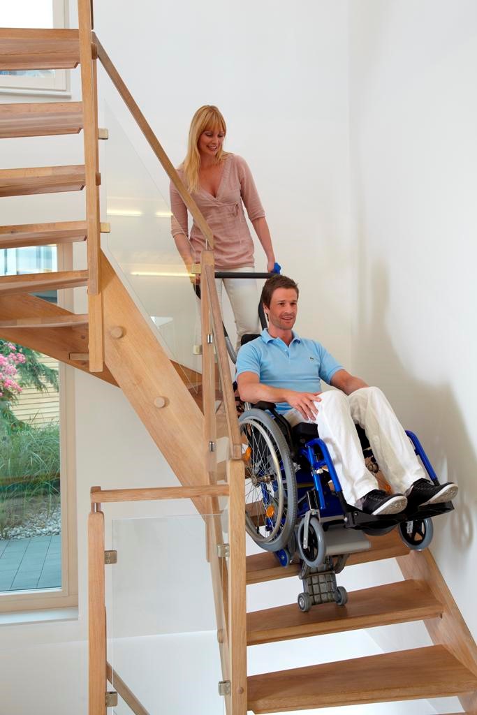 Une jeune femme manipule le monte-escalier PT Adapt sur lequel se trouve son mari. Grâce à cet appareil son mari en fauteuil roulant peut accéder aux étages de sa maison