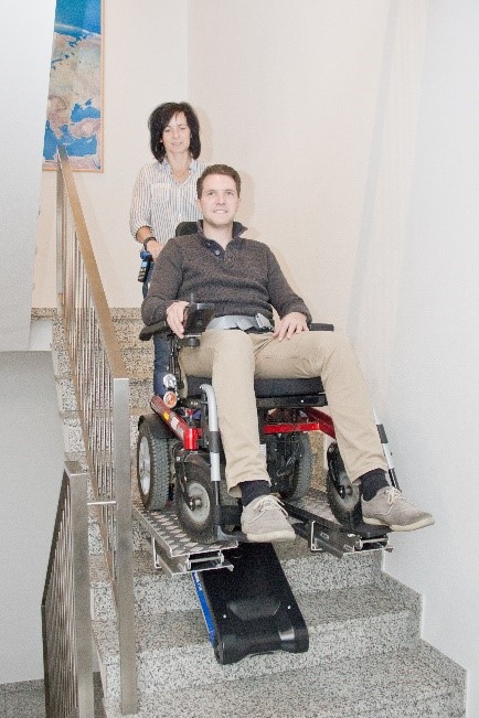 Une femme manipule un monte-escalier Liftkar PTR Long sur lequel se trouve une personne en fauteuil roulant électrique.