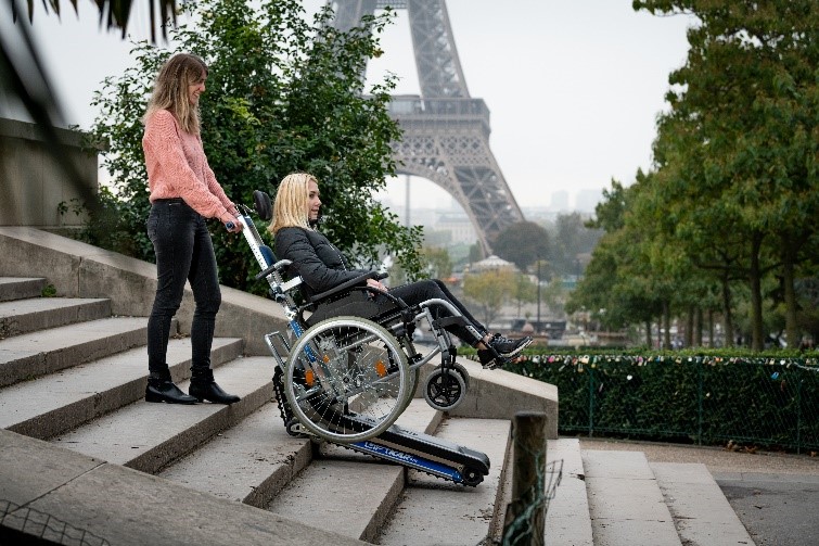 Jeune femme manipulant un monte-escaliers électrique à chenillette pour transporter dans les escaliers droits une personne en fauteuil roulant.