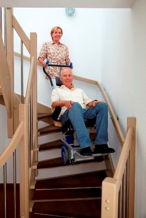 Une sénior manipule un monte-escalier électrique Liftkar PTS sur lequel se trouve son mari. Le matériel permet de monter les marches de l'escalier.