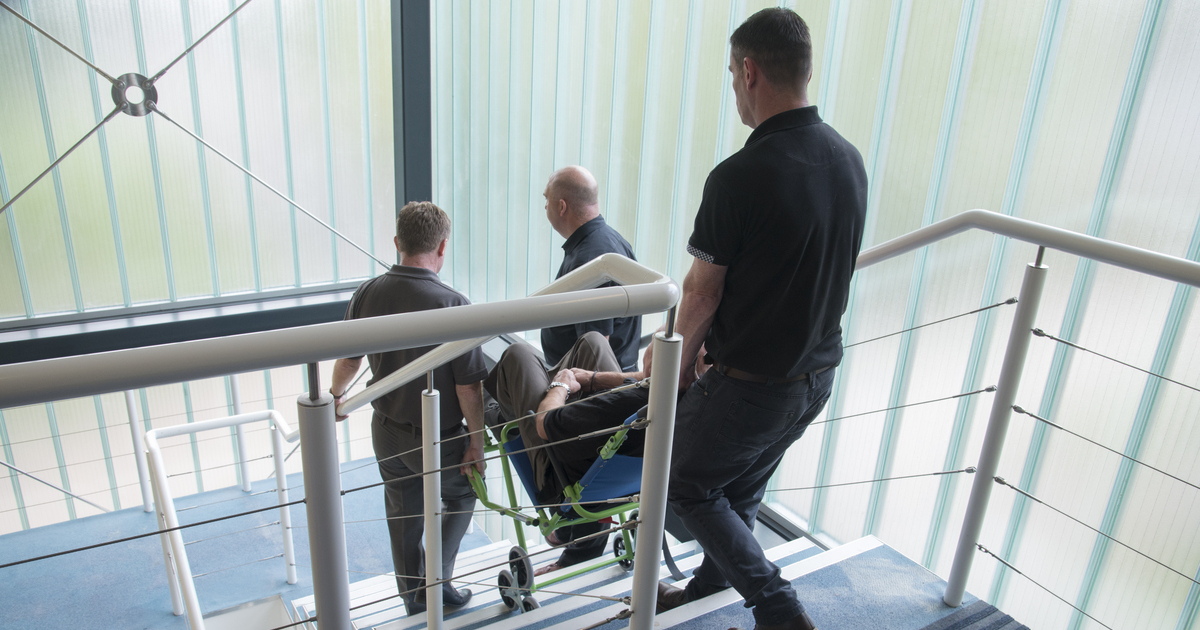 Trois aidants font descendre par les escaliers une personne à mobilité réduite sur une chaise portoir 3 roues.