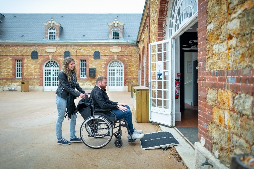 Personne en situation de handicap et son accompagnant franchissant un seuil de porte avec la rampe de voyage.