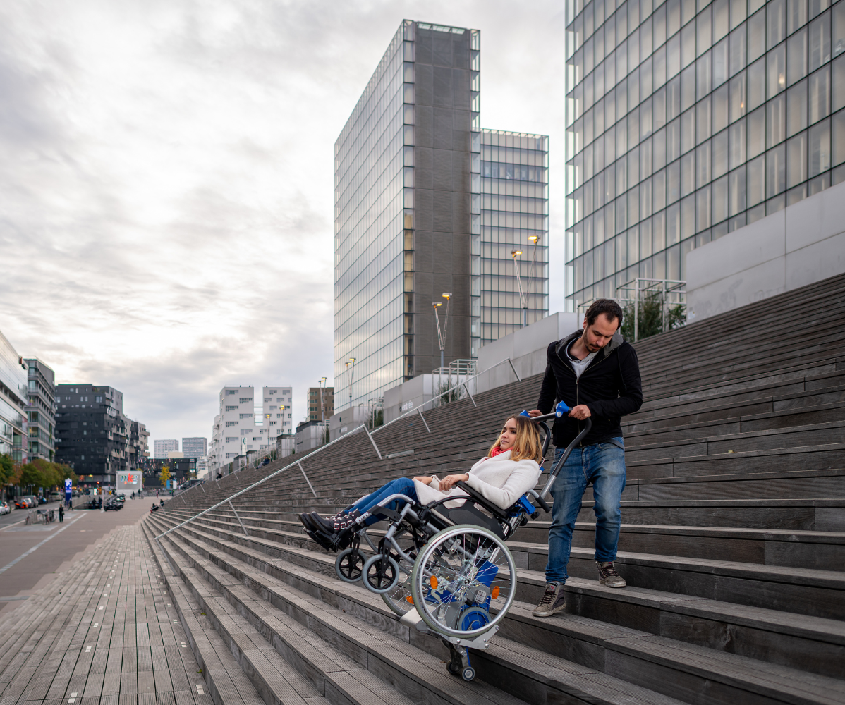 Un aidant manipule un monte-escalier Liftkar PT permettant à une personne en fauteuil roulant de monter les marches donnant accès à la Bibliothèque Nationale de France