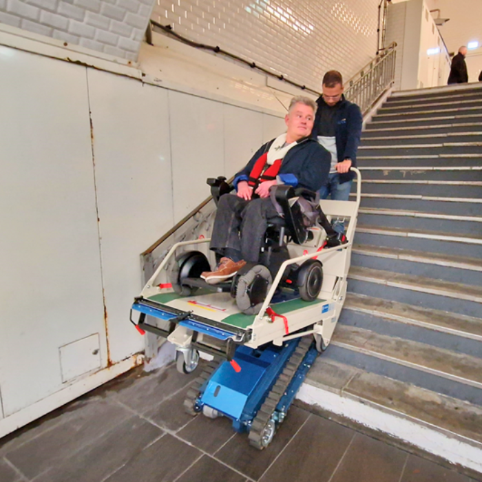 Un jeune homme manipule le monte-escaliers Atlas 230 sur lequel se trouve un homme en fauteuil roulant électrique.