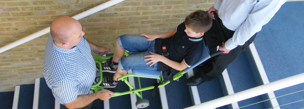 Deux aidants portent une personne à mobilité réduite dans les escaliers à l'aide de la chaise portoir 4 roues