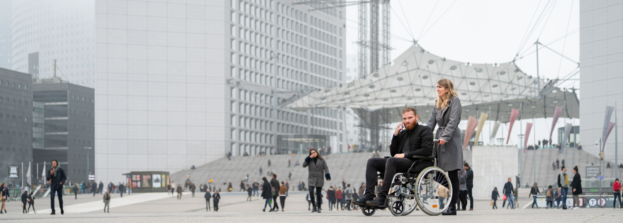 Une femme accompagne une personne en fauteuil roulant sur le parvis de la Défense