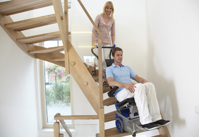 Une femme manipule un monte-escalier électrique Liftkar PTS sur lequel se trouve son mari. Le matériel permet de monter les marches de l'escalier.